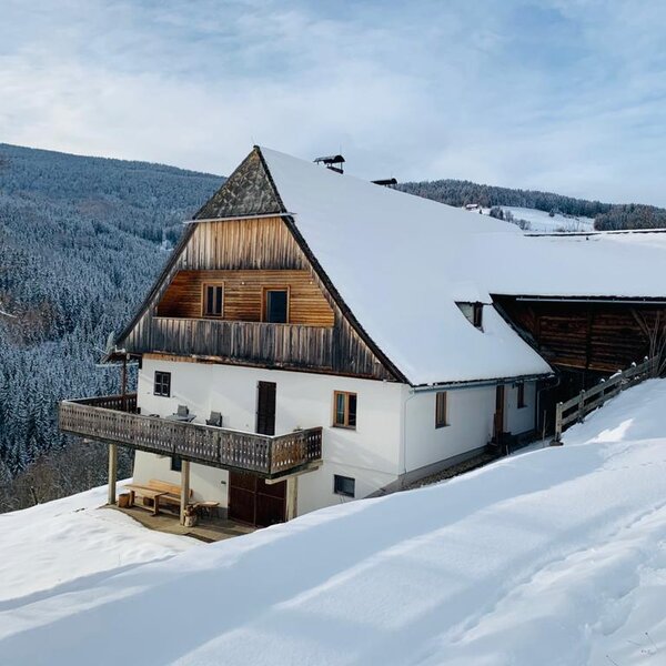 The farmhouse with a snow-covered roof and wooden balcony, set on a snowy hill surrounded by evergreen forests.