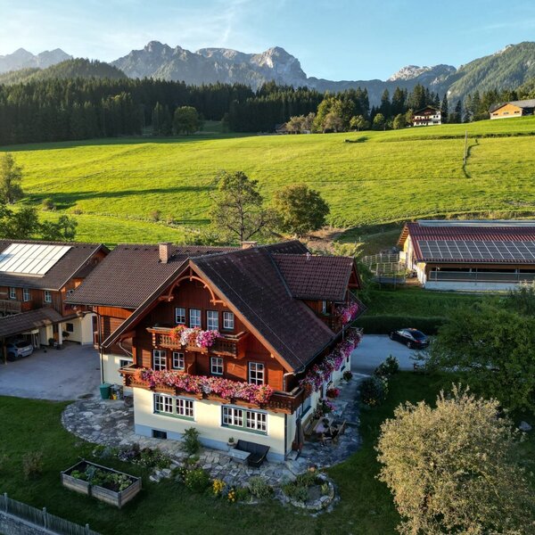 Der Bauernhof mit blumengeschmückten Balkonen, umgeben von grünen Wiesen und mit Blick auf die Berge.
