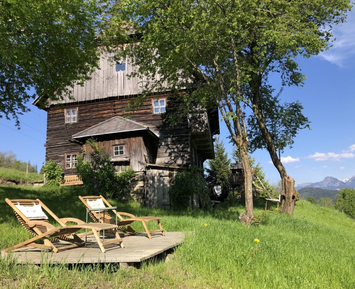 Außenansicht mit einer Holzterrasse, zwei Liegestühlen und einem Tisch, umgeben von grüner Wiese und Bäumen, mit Bergblick.