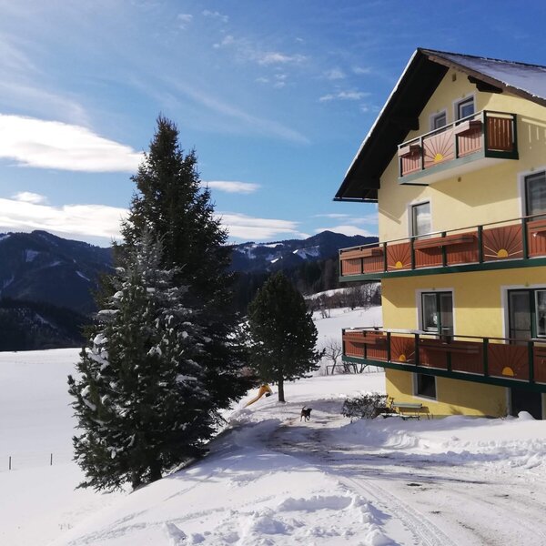 Der gelbe Bauernhof mit Balkonen, umgeben von einer schneebedeckten Landschaft, See und Bergen.