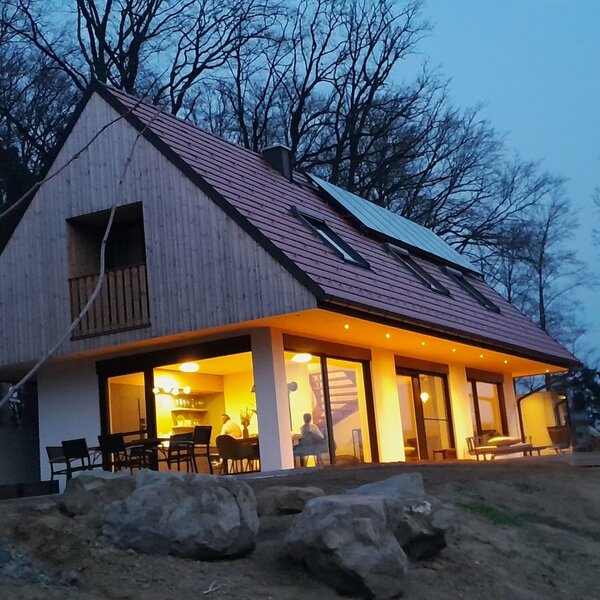 The farm house at dusk, featuring illuminated living spaces and a kitchen visible through large glass doors, with a balcony and solar panels on the roof.