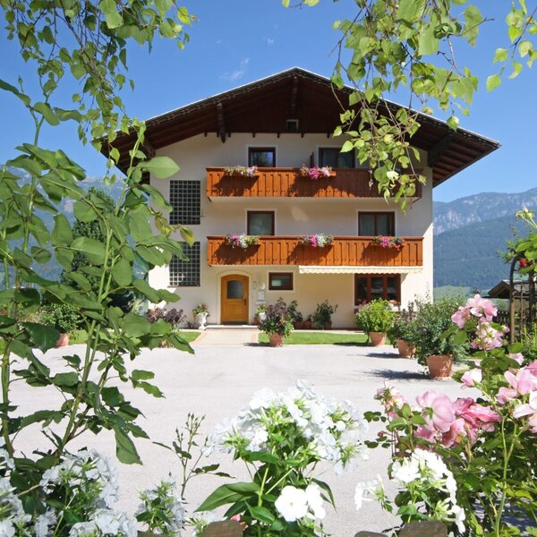 Die Außenansicht des Bauernhofs mit mehreren Balkonen, Blumenkästen und einem blühenden Garten im Vordergrund, mit Bergblick im Hintergrund.