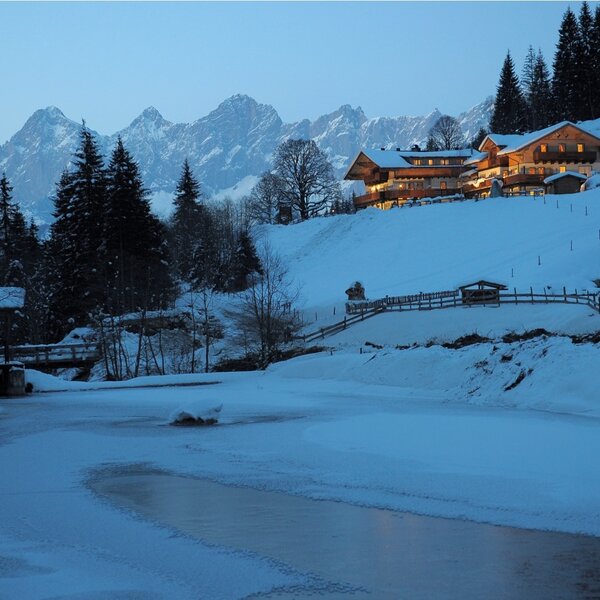 Der verschneite Bauernhof mit beleuchteten Fenstern in den Bergen und einem teilweise zugefrorenen Gewässer im Winter.