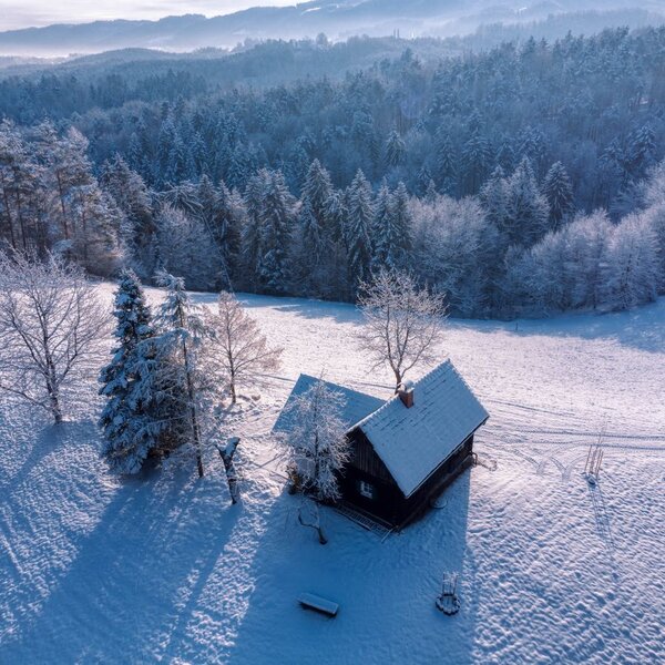 The farmhouse in a snow-covered meadow with a forest and hills in the background.