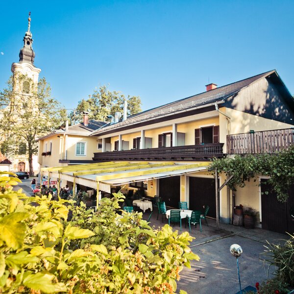 The farmhouse exterior offers an outdoor dining area with tables and chairs under a canopy and features upper-floor balconies, with a church steeple visible in the background.
