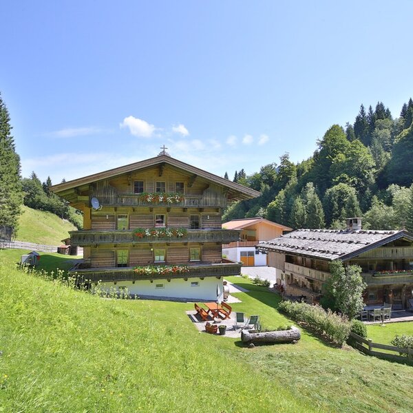 The farmhouse exterior, featuring traditional wooden construction, balconies with flower boxes, and an outdoor seating area on a grassy slope.
