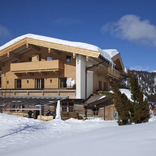 The exterior of the Farm House in winter, featuring wooden construction, snow on the roof, balconies, a covered outdoor terrace, and a mountain backdrop.