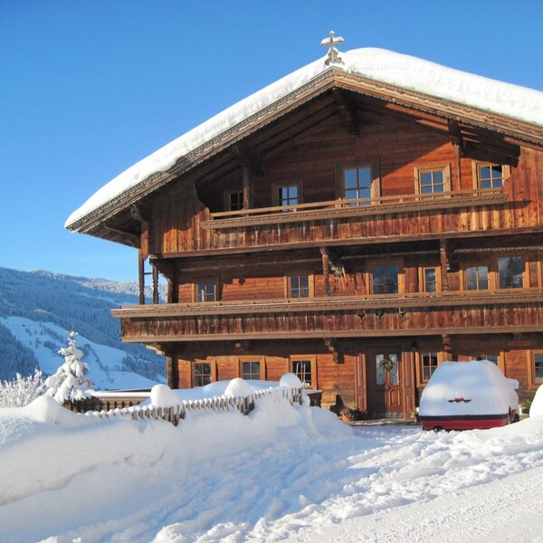 The traditional wooden exterior of the Farm House, with snow on the roof and balconies, set against a winter mountain landscape.