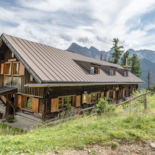 Die Alm, eine Holzhütte mit Metallblechdach und Fenstern mit Holzläden, gelegen in einer bergigen Landschaft.