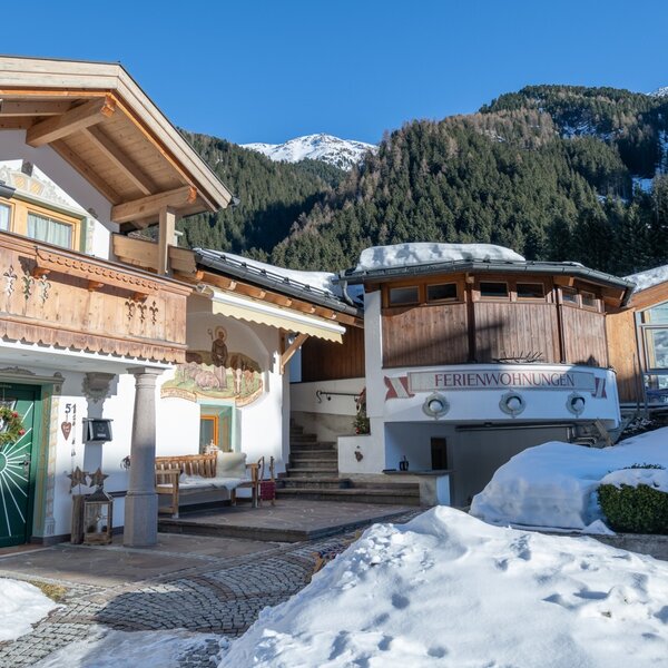 Außenansicht des Bauernhofs mit traditionellen Holzelementen, dem Schild „Ferienwohnungen“ und schneebedeckter Umgebung vor den Bergen.