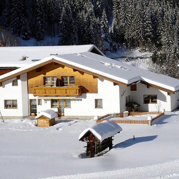 The Farm House "Bligg" covered in snow, featuring a balcony, a children's swing set, and surrounded by a snowy landscape and pine forests.