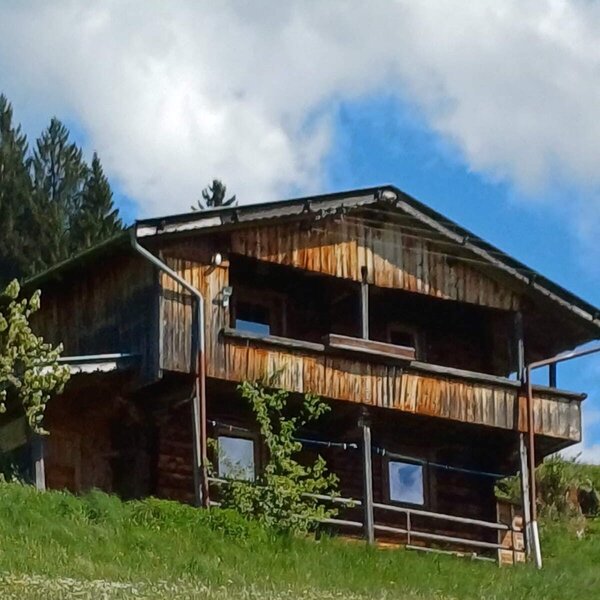 The wooden Alpine Hut features multiple balconies and is situated on a green, flowered slope.