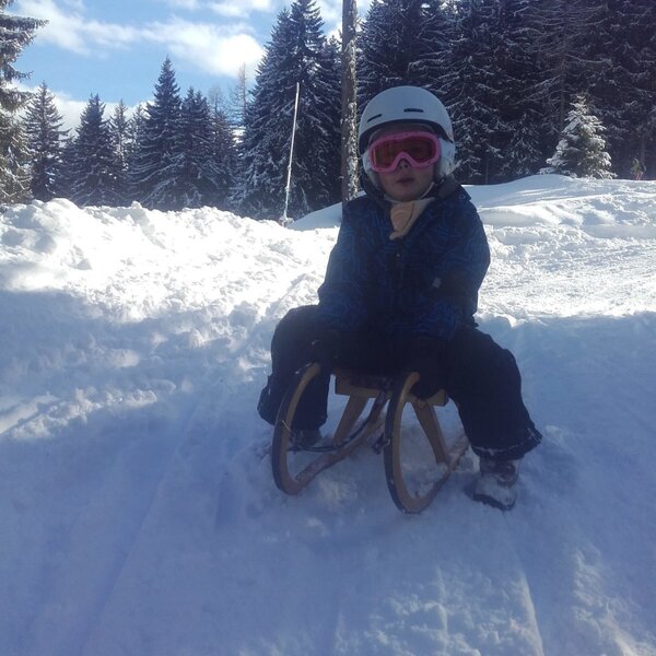Ein Kind auf einem Holzschlitten beim Rodeln in der verschneiten Winterlandschaft rund um den Bauernhof.
