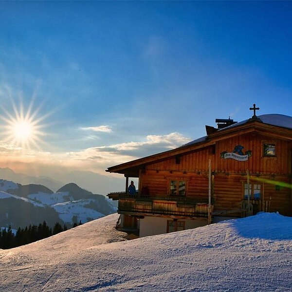 The wooden Alpine Hut features a balcony and is situated on a snowy mountain slope, offering expansive views under a bright sky.