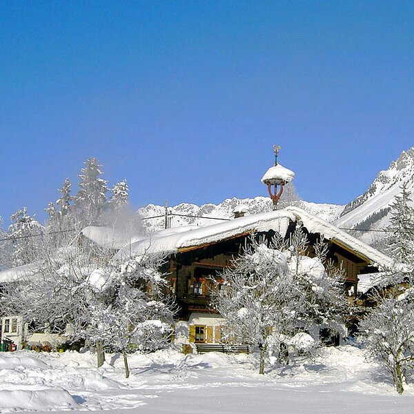 Der schneebedeckte Bauernhof vor einer winterlichen Berglandschaft unter blauem Himmel.