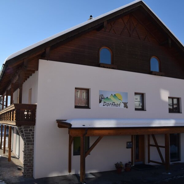 Außenansicht des Bauernhofs "Dorfhof" mit verschneitem Dach und traditionellem Holzbalkon.