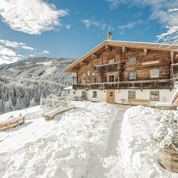 Der Bauernhof in verschneiter Winterlandschaft mit traditioneller Holzfassade, Balkonen und einem Zugangsweg.