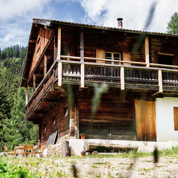 Außenansicht der Alm mit dem sonnigen Balkon, einem Sitzbereich im Garten und einem Brunnen.