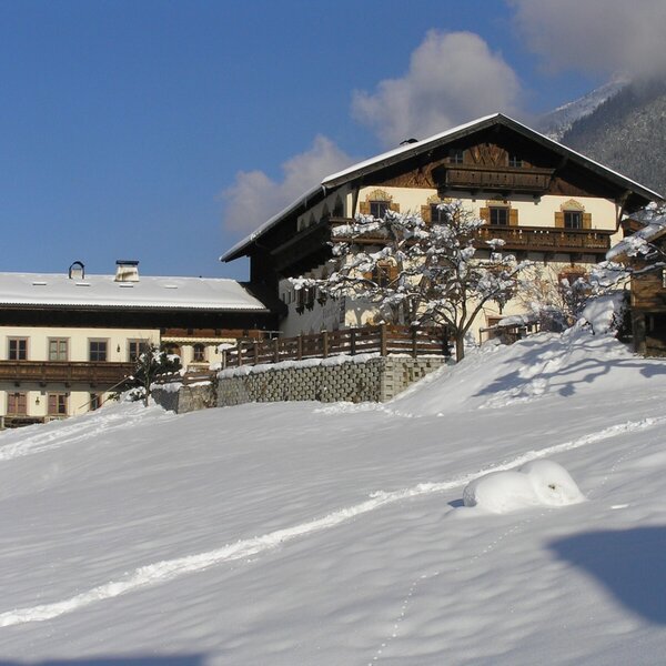 The Farm House exterior in winter, featuring traditional architecture with balconies, surrounded by snow-covered grounds and distant mountains.