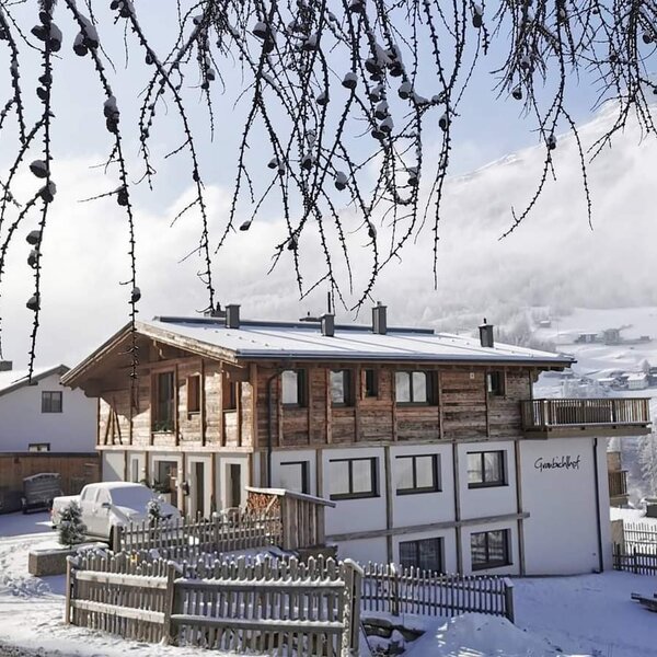 The snow-covered Farm House, featuring traditional wood and white architecture, a balcony, and mountains visible in the distance.