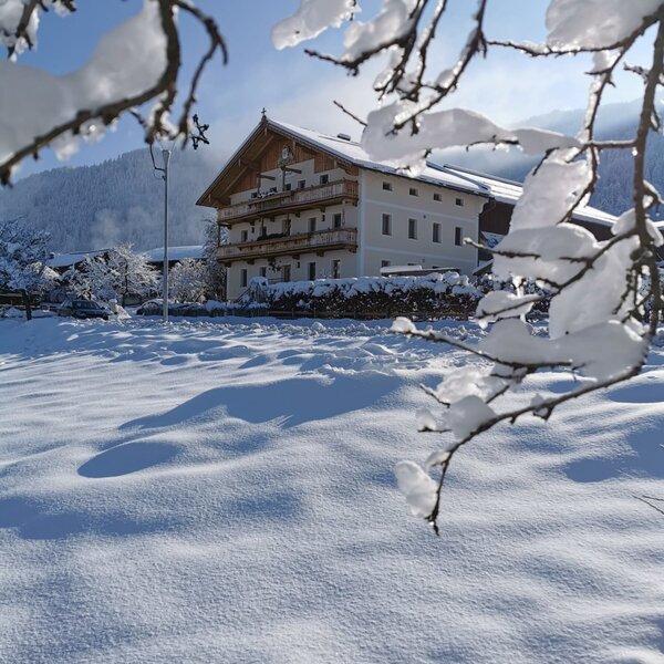 Der Bauernhof im Winter, mit schneebedeckten Balkonen und umgeben von Schnee vor einer Bergkulisse.