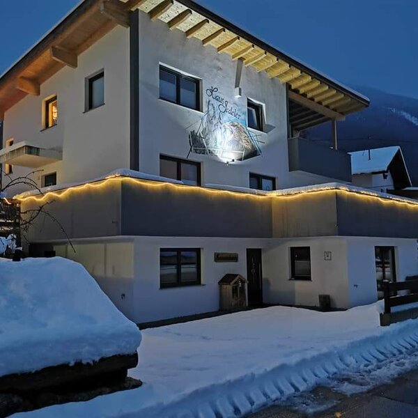 Die winterliche Außenansicht des Bauernhofs mit Schnee und dekorativer Beleuchtung bei Dunkelheit und Bergblick.