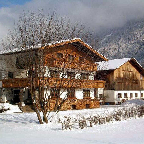 The Farm House exterior during winter, showcasing traditional alpine architecture, wooden balconies, and a snow-covered landscape with mountains in the background.