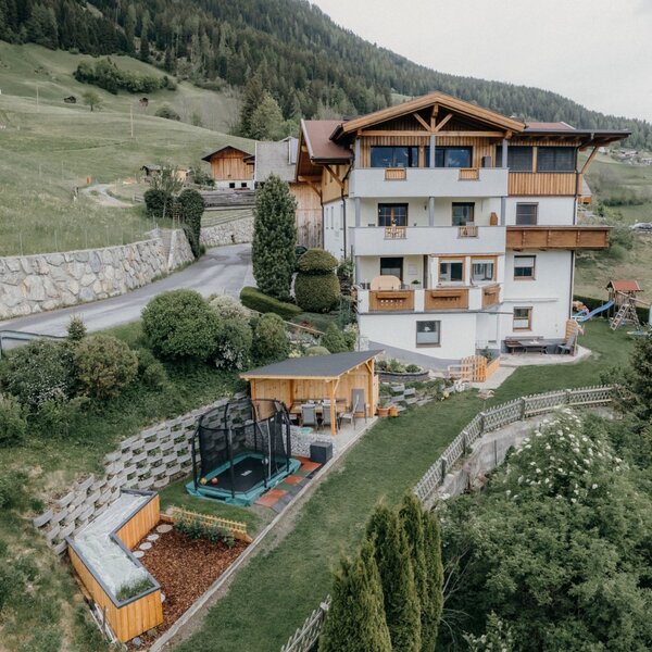 The Farm House features a garden with a trampoline, a children's playground, a covered outdoor seating area, and multiple balconies, set on a hillside with mountain views.