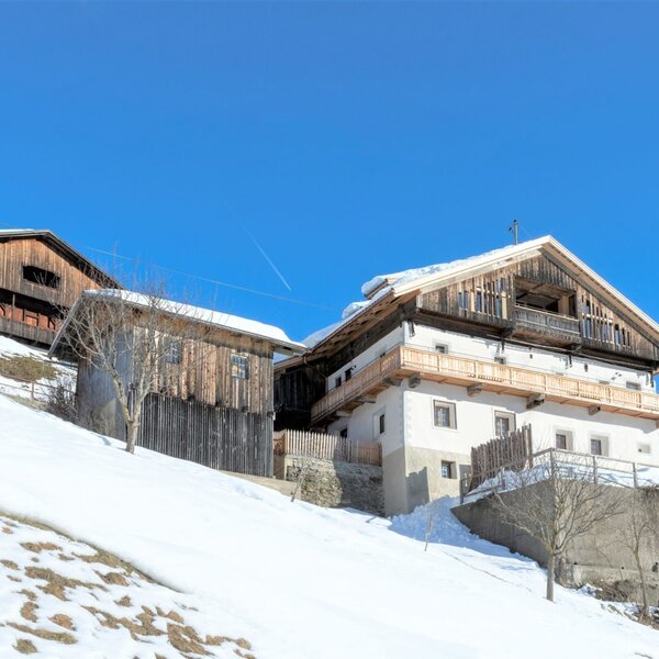The farm house with white walls, wooden balconies, and a gabled roof is situated on a snow-covered hillside, alongside other wooden buildings.