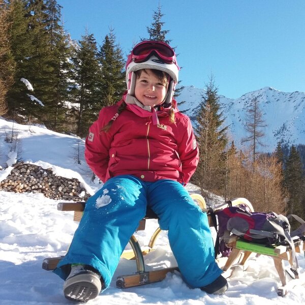 Winterliche Familienaktivitäten am Bauernhof: Ein Kind auf einem Schlitten im Schnee vor der Berglandschaft.