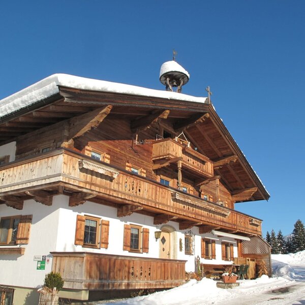 The Farm House exterior during winter, featuring traditional wooden balconies, a rooftop bell, and snow-covered surroundings.