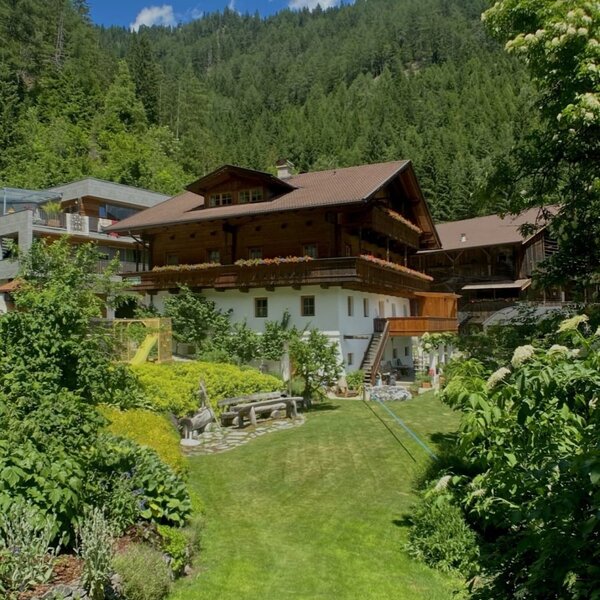The Farm House exterior with traditional architecture, balconies, a green lawn, and garden, set against forested mountains.