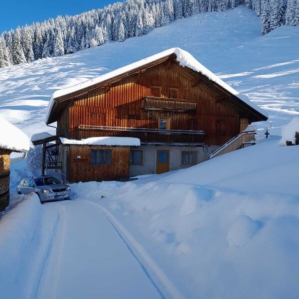 The wooden Alpine Hut in a snow-covered mountain landscape, featuring balconies and a snow-cleared driveway.
