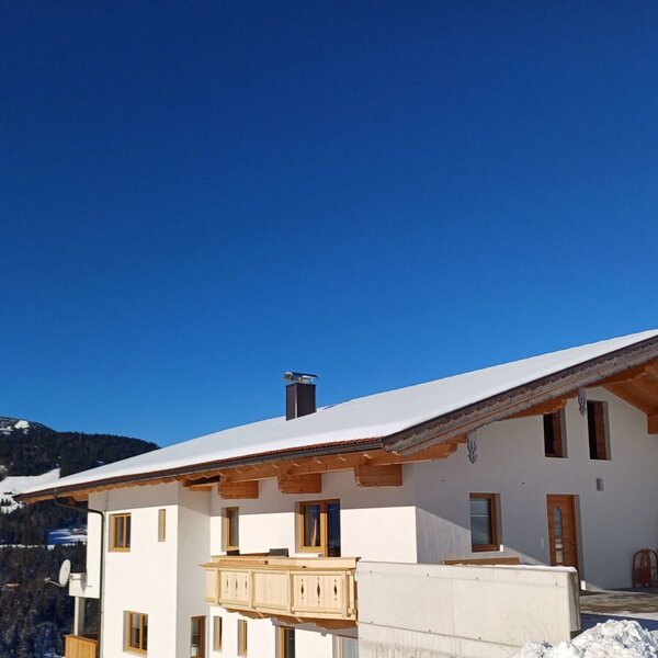Der Bauernhof von außen mit verschneitem Dach, Holzbalkon und Bergblick im Winter.