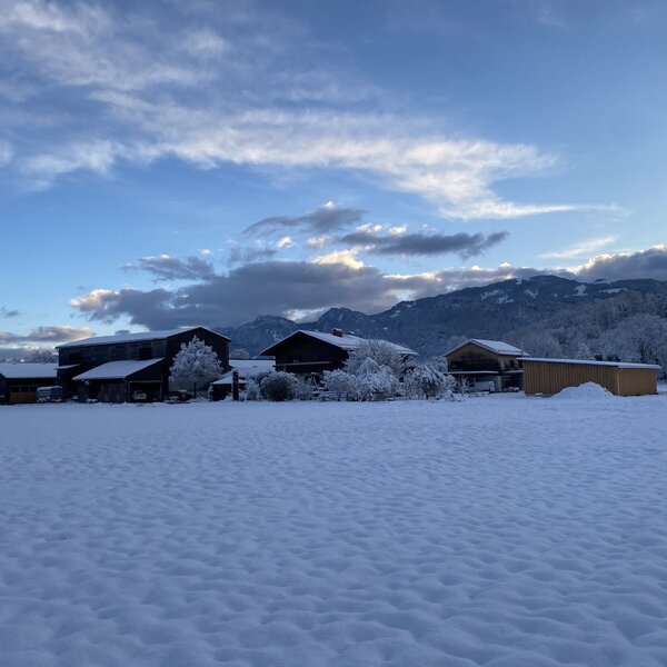Der Bauernhof in verschneiter Winterlandschaft mit Feldern und Bergen im Hintergrund.