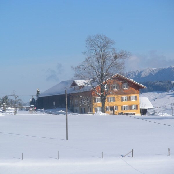 Außenansicht des Bauernhofs im Winter, umgeben von schneebedeckten Feldern und Bergen unter blauem Himmel.