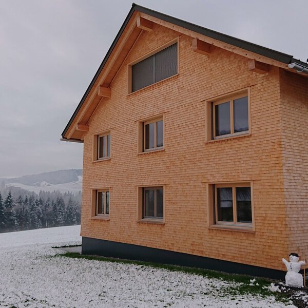 The exterior of the newly built Farm House, featuring a light wooden facade, multiple windows, and a snow-covered landscape with evergreen trees.