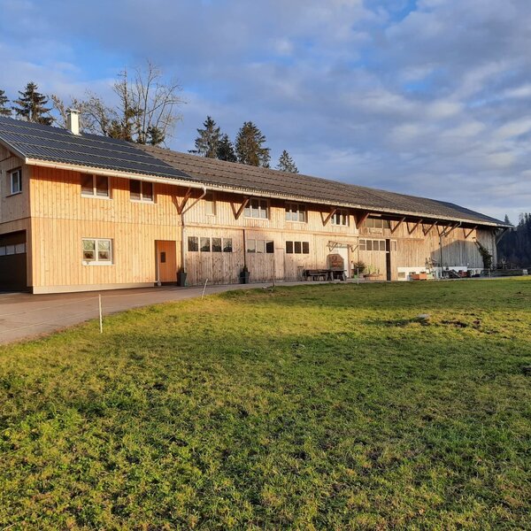 Der Bauernhof mit Holzfassade, Solarmodulen auf dem Dach und einer Rasenfläche vor dem Gebäude.